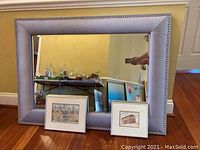 Photo of two signed framed drawings leaning against a large fabric framed mirror with silver nailhead trim.