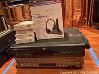 Four boxed itek Bluetooth Track & Find 2-pack devices stacked next to boxed RadioShack Wireless Headphones atop two video players: Hitachi DVP305U DVD player (black) and Samsung DVD-V3500 DVD/VHS player (silver/gray).