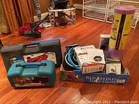 Overall lot overview showing hydraulic jack case, Makita sander case, exercise mat, heating pads, Pilates DVDs, Fitbit band, and Chirp wheels arranged on wooden floor.