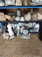 Wide shot of multiple stone lawn figures including owls, birds, and rabbits stacked on two metal shelves and floor.