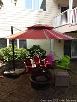 Photo of red patio umbrella open with red, pink, and green Adirondack chairs and fire pit beneath on brick patio.