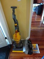 Full upright Dyson vacuum with yellow and gray finish, with home cleaning kit box and manual on floor beside it