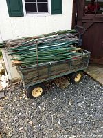 Photo of rusty metal garden wagon loaded with metal fence posts, wooden stakes, and bamboo rods outside against shed wall on gravel surface.