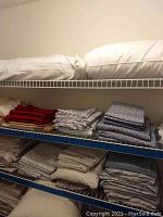 White bed pillows on top shelf, folded sheets underneath including red and patterned gray sets, beige, and gray several folded sheet sets on bottom shelves