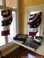 Four patriotic red, white, and blue star-spangled window swags hanging on windows and door with a white table below holding crafting tools and decorations.