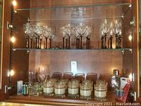 A set of various glasses including wine glasses with black stems on top shelf, shot glasses with frosted white bands on middle shelf, along with a glass flask, vacuum stoppers, and a corkscrew on lower shelf inside a display cabinet.