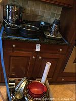 Two Berndes black cast pots with lids on countertop, stainless steel Cookworks steamer pot, roasting pan bottom, graters, and mixing bowls in cabinet drawer below.