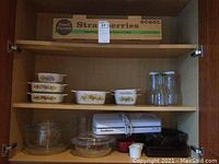 Photo of wooden shelf with three shelves showing Corning Ware glass dishes with vegetable decals, Food Saver Vac 1050 on bottom shelf, clear glass bowls, and tall glass bottles, also some dark items possibly flatware tray.
