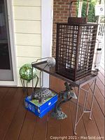 Overall view of the lot showing all five items: gazing ball on metal stand, turtle statue on box, fairy statue on floor, metal plant stand, and brown metal lantern on top of the stand.