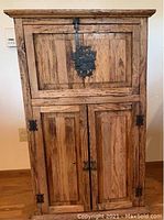 Front-facing image of a rustic solid wood storage cabinet with two compartments. The cabinet has a medium natural wood finish with visible grain and black metal hardware including a latch on top compartment and handles on lower double doors.