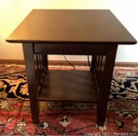 Front view of wooden side table on a patterned rug, showing square shape, flat top, bottom shelf, and legs.