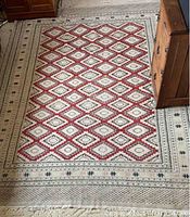 Full view of the oriental wool carpet laid on a floor next to furniture showing the geometric diamond pattern in red, cream and black with wide cream border and fringe edges.