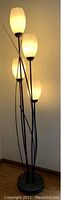Image showing a tall metal floor lamp with four frosted glass or plastic lampshades arranged vertically, emitting warm light.