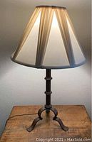 Wrought iron table lamp illuminated on wooden surface showing pleated fabric lamp shade and tripod iron base.
