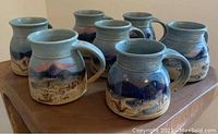Seven handmade ceramic mugs with painted mountain landscapes and blue glaze details, displayed on a wooden surface.