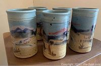 Photo showing 5 Fire Hole pottery tumblers with painted mountain and desert landscape scenes. Tumblers have a blue sky gradient and earthy bottom with painted shrubs.