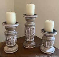 Photo showing three carved wooden candlesticks each topped with an off-white pillar candle. Candlesticks have whitewashed carved leaf and geometric designs with a rustic finish. Varying heights from 5 to 7 inches.