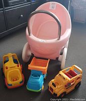 Pink Little Tikes baby carriage with three toy trucks including a yellow bus shape sorter and a blue and orange dump truck visible in front
