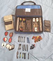 Open blue and brown fabric carrying case showing steel and soft tip darts neatly arranged inside with additional shafts, flights, and 12 spare pins spread around it. Also includes a smaller brown pouch and a metal bottle opener tool.