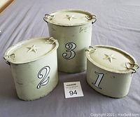 Three white tin canisters numbered 1, 2, and 3 on a cloth surface with lids on.
