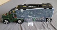 Side view of green dinosaur carrier truck showing transparent compartment with multiple toy cars and dinosaur figures inside.