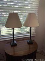 Pair of lamps on a wooden table in front of a window with blinds. Both lamps have black metal bases and beige fabric shades.