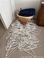 Shows the woven laundry basket partially filled with folded wool blankets with tartan patterns, accompanied by white plastic hangers scattered on carpet