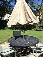 Photo showing round heavy metal patio table with beige umbrella in the center and metal base visible underneath. Surroundings include grass and patio surface. Scratches visible on tabletop.