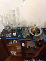 Wide shot of assorted glassware set on a dark surface including wine glasses, beer mugs, pitcher, decanters, perfume bottle, silver plated tray and butter tray