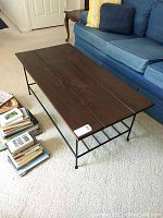 Coffee table with three dark stained wooden planks on top and black metal frame and legs in a living room setting next to a blue sofa.