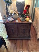 Front view showing the wooden cabinet style end table with two doors, bronze tone hardware, and various decorative items on top (not included in lot).