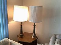 Two matching mid-century modern style lamps with burnished brass bases and white fabric shades placed together on an octagonal wooden table beside a couch.
