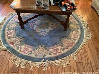 Full view of round blue and cream carved area rug with floral center medallion, fringe on edges, placed on wood floor under table with items on top
