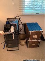 Overview of metal cart stacked with phones, pencil sharpeners, boxes of legal size hanging file folders, waste basket next to boxes