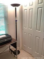 Black metal torchiere floor lamp with upward facing bowl-shaped shade beside white doors with window blinds in background.