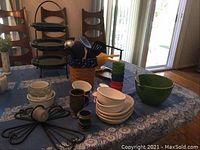 Wide view of kitchen utensils in holder, stacking cups, bowls, plates and plate stand on table with blue tablecloth