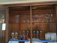 View of multiple crystal stemware glasses arranged inside a wooden display cabinet, showing various sizes and etched designs, including four multicolored cordial glasses at bottom center.