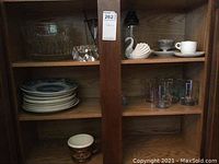 Shelf with stacked white plates, clear glass serving bowls, faceted glass candle holders, swan figurine, teacups and glasses