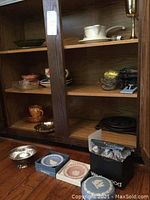 Overall lot showing glass and metal trivets, plates, bowls inside wooden cabinet and three Wedgwood boxed dishes on floor
