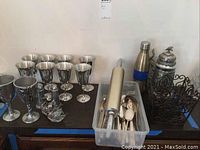 Full view of the lot on a black table, showing metal goblets, baskets, water bottle, stein, rolling pin, flatware in container, and salt cellars.