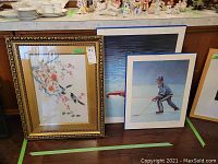 Framed Asian embroidery art and two sports prints leaning against a wall on the floor with other items in background