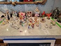 Wide image showing various ceramic and porcelain figures, including men and women figurines, animal shapes, and a piggy bank on a marble surface with ornate background.