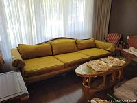 Full view of the long mustard yellow velvet sofa with wood base and feet, situated in a living room with natural light coming through sheer curtains.