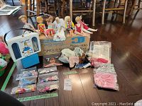 Wide shot of the seven vintage Barbie dolls displayed seated on a cardboard box labeled 1960's-1970's collectibles, with doll clothing, grouped Barbie shoes bagged separately, and other doll accessories including a Lollipop play case and doll hangers.