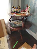 Full view of the vintage wooden two-tiered side table with various decorative porcelain figurines on top and a decorative plate on lower shelf.