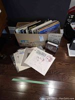 Box containing more than 20 vintage 78rpm LP records and paper sleeves, curated for variety of genres and artists.