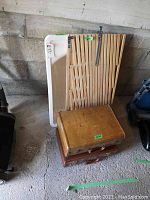 Photo showing two stacked vintage leather suitcases, wooden expandable baby gate behind, and a white plastic baby bath tub leaning against the wall.