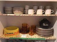 Shelves with stacked light colored stoneware cups, saucers, side plates, and dinner plates, along with amber colored glass tea cups and saucers, a yellow oversized dog food bowl, and a black ceramic Planters nuts bowl with lid.