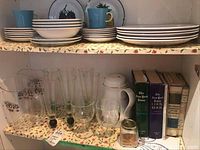 Shelf with stacked Mikasa plates, bowls, cups, clear cocktail glasses, plastic Coors pitchers, thermal coffee carafe, vintage cookbooks