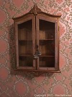 Front view of ornate wooden wall shelf with glass doors closed and key lock.